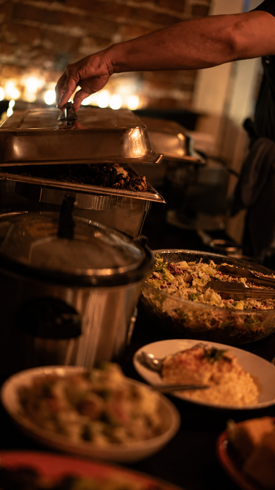 A buffet-style food scene with a person's arm reaching over a chafing dish, with plated meals and cooking pots in a warm indoor setting.