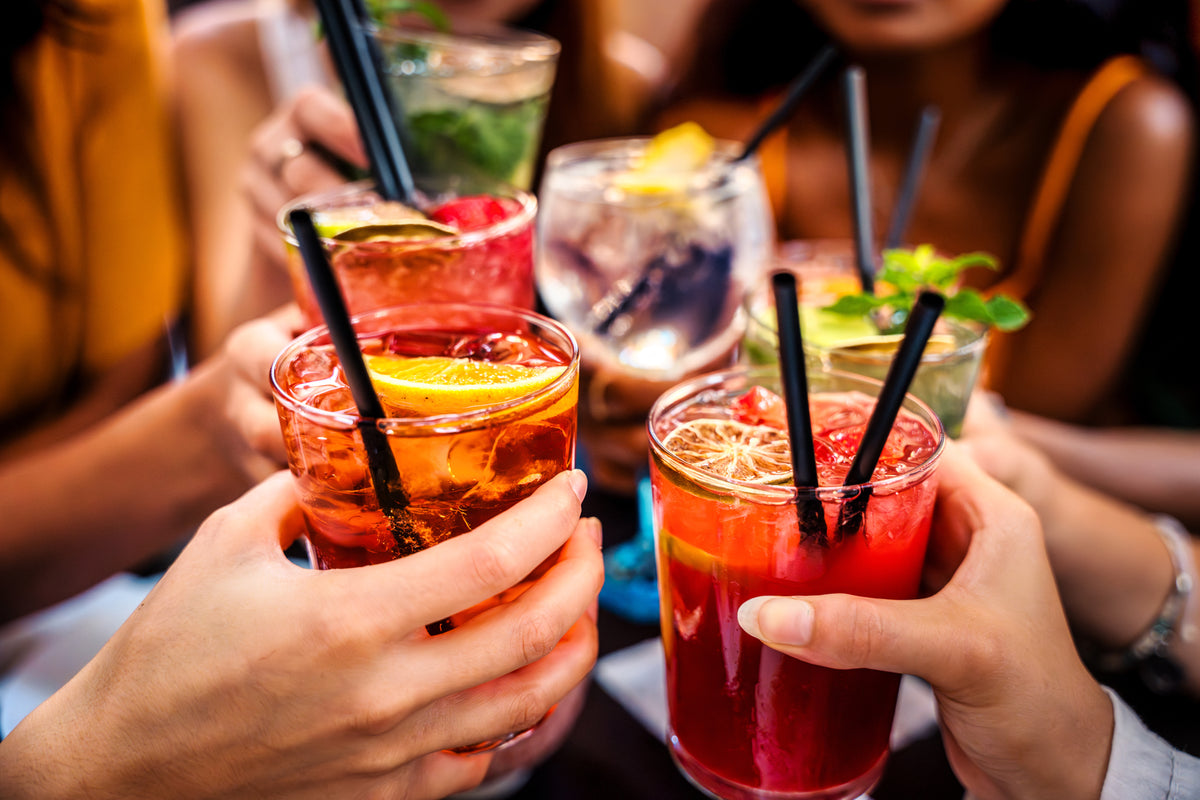 Several colorful cocktails in clear glasses with black straws are being held up in a close-up toast scene. Hands are visible holding the drinks, and blurred people and a real social environment appear in the background.